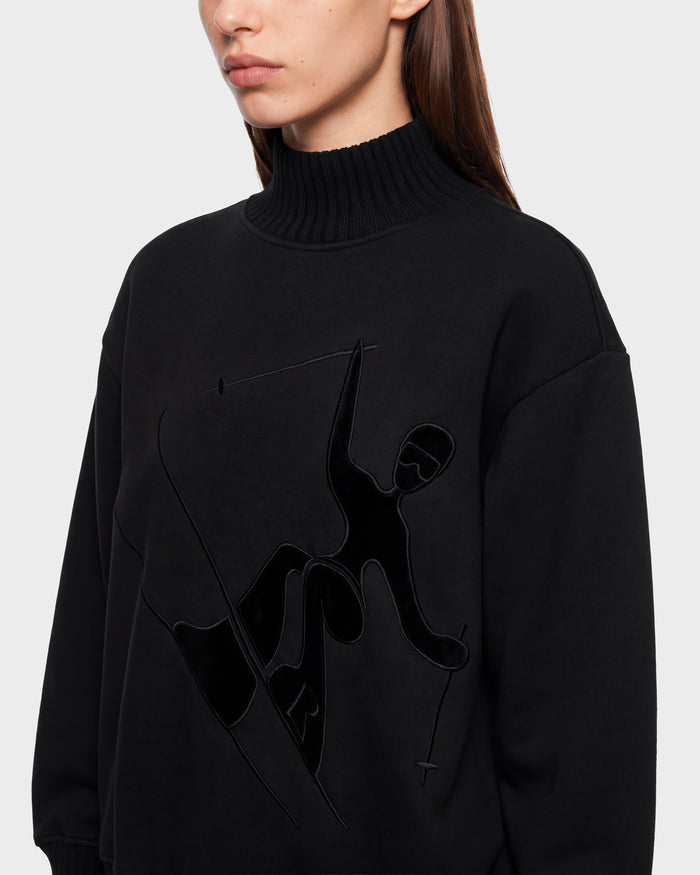 A woman wearing the Rudsak JOYCE E - 8526538 Black turtleneck sweatshirt, which features an embossed graphic of a skier holding poles on the front. She is shown from the shoulders up, facing slightly to the left.
