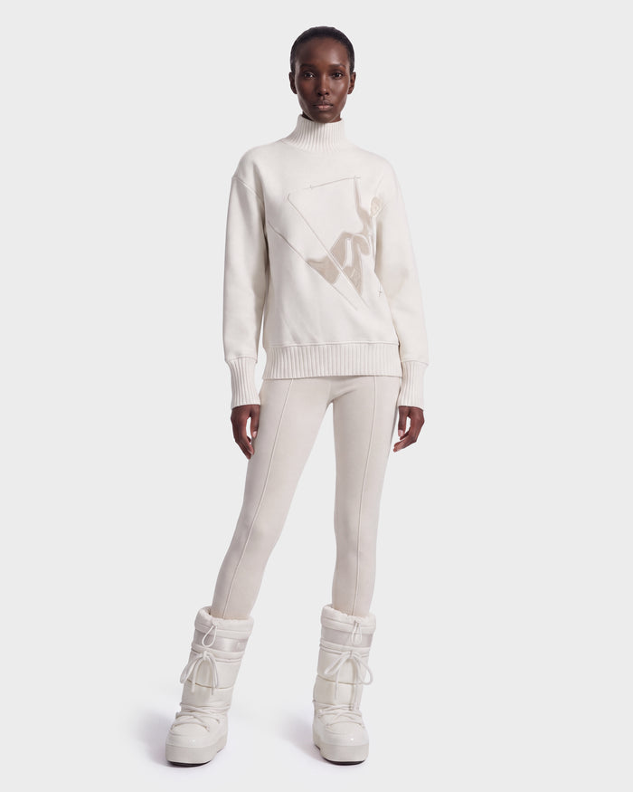A person stands facing forward against a plain white background, wearing the JOYCE E - 8526538 Vanilla tall, padded boots by Rudsak, paired with an ivory turtleneck sweater and matching fitted pants.