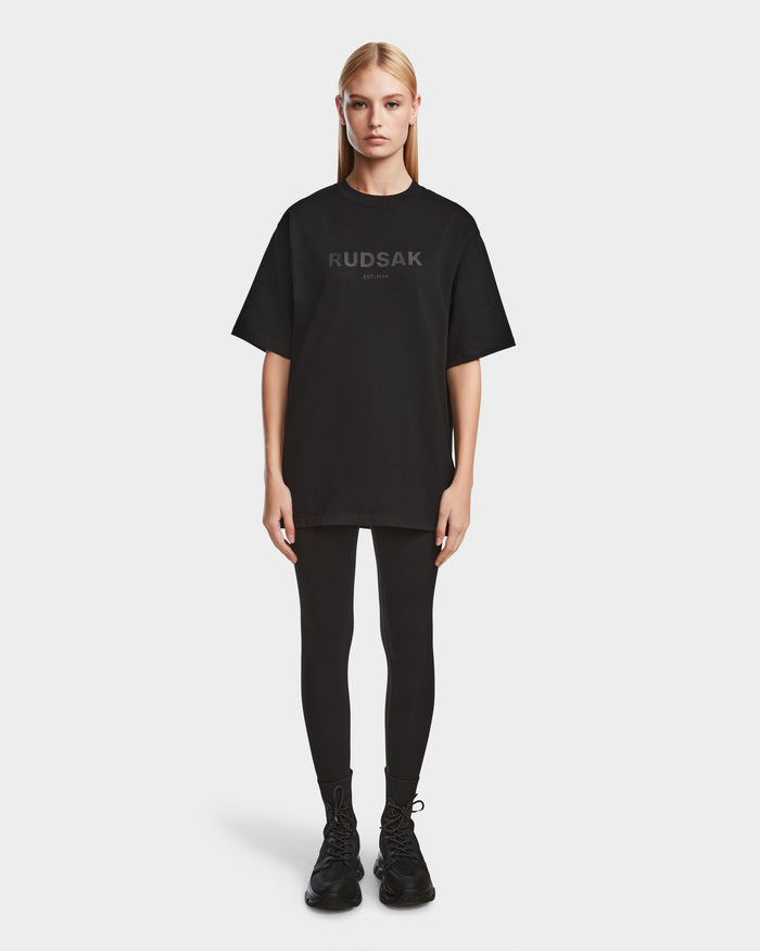 A woman with straight blonde hair wears Rudsaks KARTER - 9524008 Black W, a relaxed-fit oversized unisex premium cotton T-shirt with RUDSAK and MONTREAL on the front, paired with black leggings and shoes against a white background.
