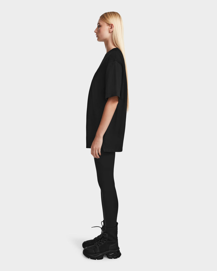 A person with long blonde hair stands in profile, wearing the Rudsak KARTER - 9524008 Black W relaxed fit oversized unisex T-shirt, paired with black leggings and chunky black shoes against a plain light background.