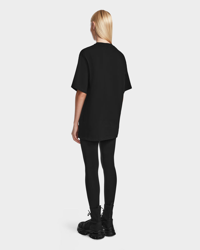 A person with straight blonde hair faces away, wearing the Rudsak KARTER - 9524008 Black W oversized unisex T-shirt, paired with black leggings and sneakers against a plain light background.