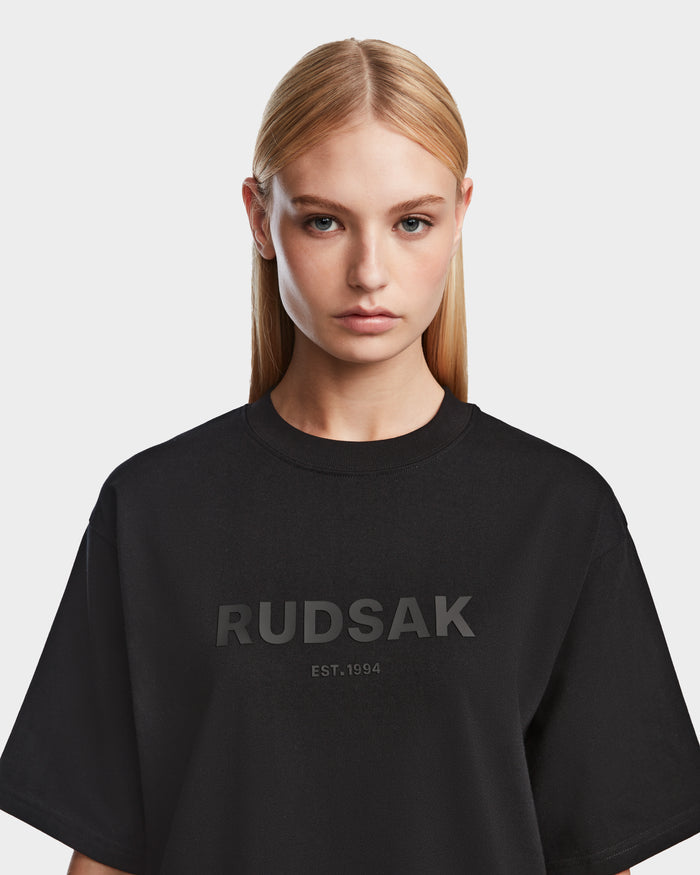 A woman with straight blonde hair wears the KARTER - 9524008 Black W relaxed fit T-shirt by Rudsak, featuring RUDSAK and EST. 1994 on the front, standing against a plain white background and looking at the camera.