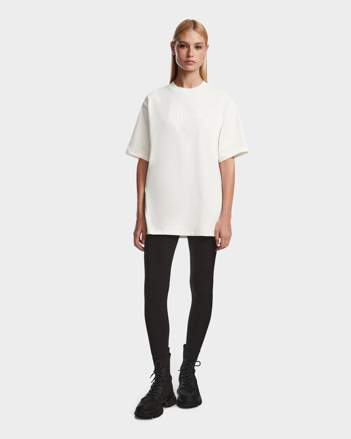 A woman with straight blonde hair wears the Rudsak KARTER - 9524008 Iceland W premium cotton T-shirt, black leggings, and black lace-up boots while standing against a plain white background.