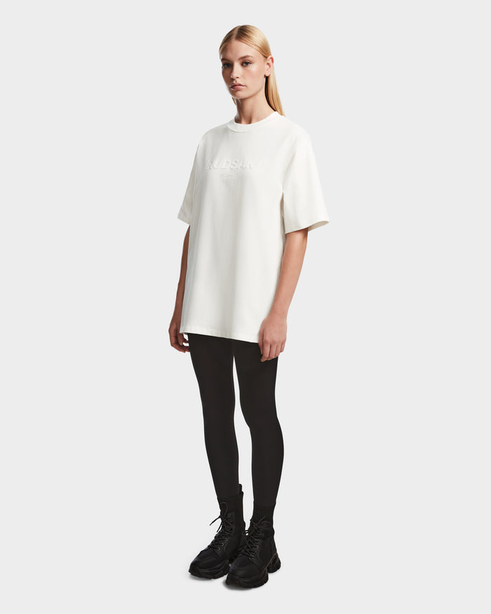 A person with long blonde hair stands against a plain background, wearing the Rudsak KARTER - 9524008 Iceland W T-shirt, black leggings, and black lace-up boots.
