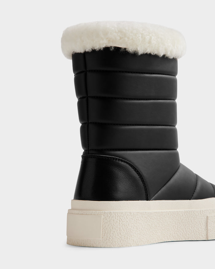 A close-up of the Rudsak KATHIE - L225516 Black women’s mid boot features a quilted design, thick white sole, and fluffy faux fur trim at the top, shown against a plain light background.