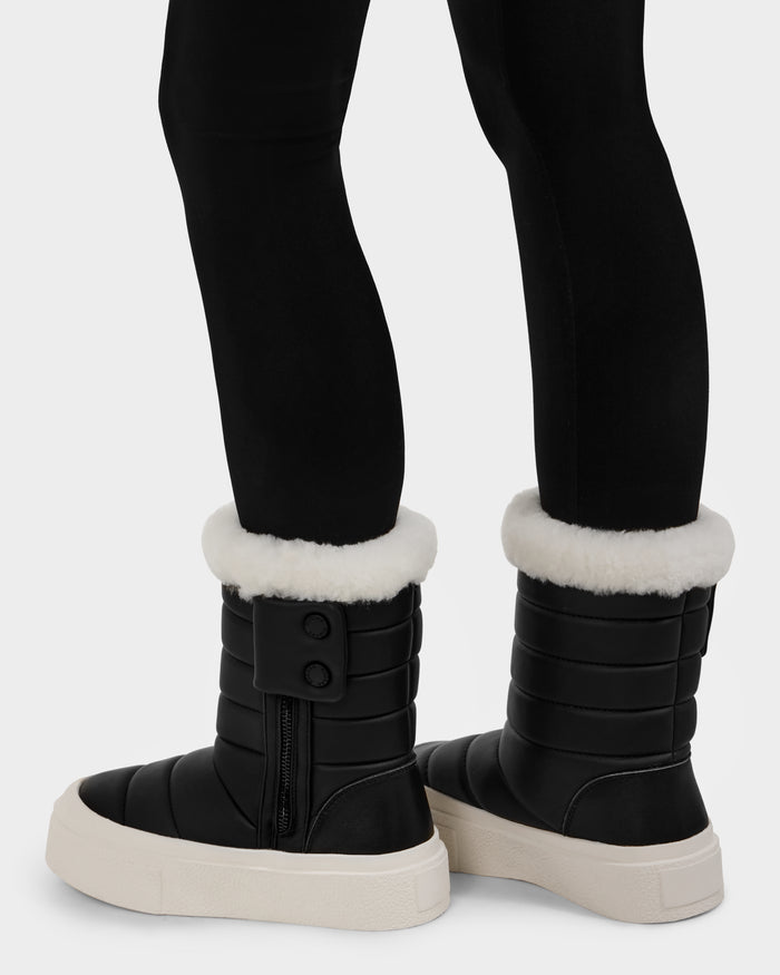 A person wearing Rudsak KATHIE - L225516 Black women’s winter boots with white faux fur cuffs, thick white soles, side zippers, and two decorative buttons, paired with black leggings.
