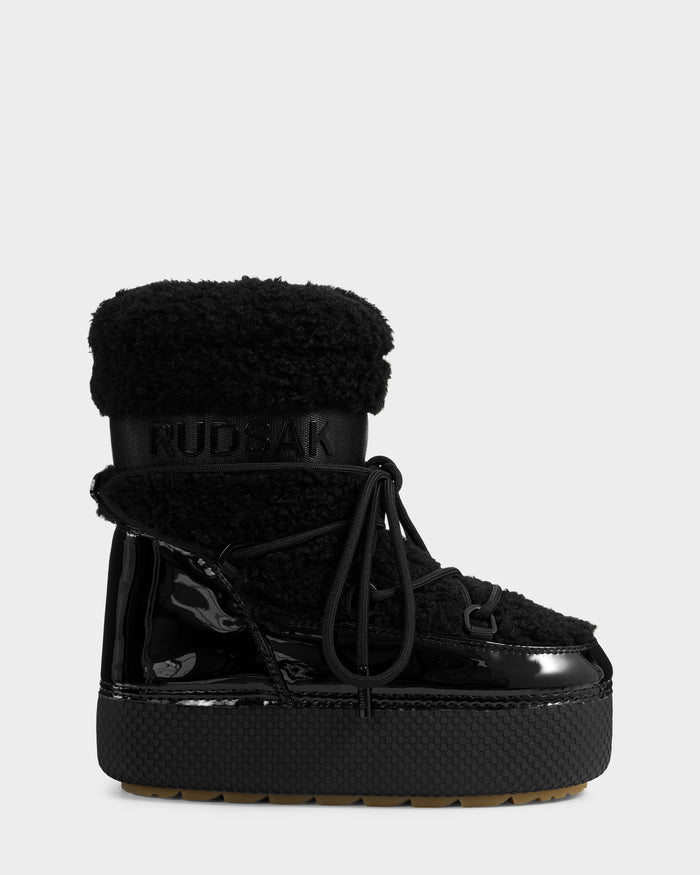 A single KATHY Y - 1224500 Black winter boot by Rudsak features a shiny patent finish, textured sole, thick laces, faux fur upper cuff, and an embossed Rudsak logo on the shaft. The background is plain light gray.