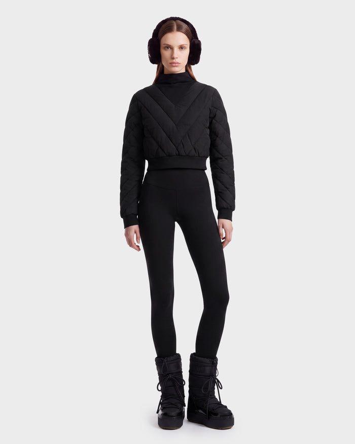 A person stands facing forward against a plain light background, wearing the Rudsak KATYA - 8526531 Black quilted down sweater, complemented by black earmuffs, black leggings, and black winter boots.
