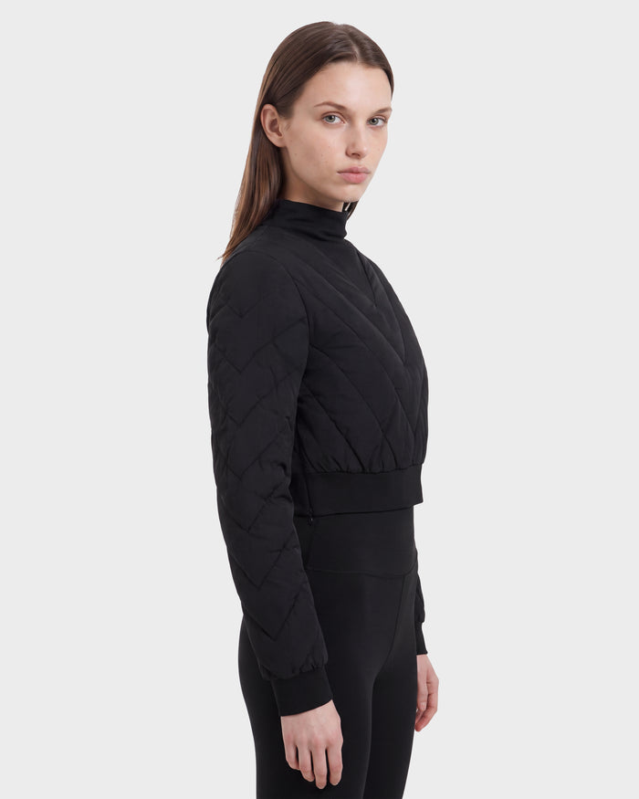 A person with straight brown hair is wearing the Rudsak KATYA - 8526531 Black quilted down jacket and black pants, standing sideways against a plain light gray background.