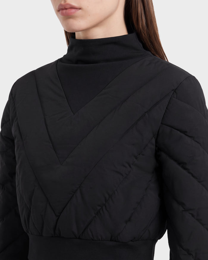 A woman is seen from the upper body up, partially revealing her face, styled in an effortlessly chic après-ski look. She wears the Rudsak KATYA - 8526531 Black jacket—a quilted, V-patterned down-insulated piece—layered over a high-neck black top, set against a plain backdrop.