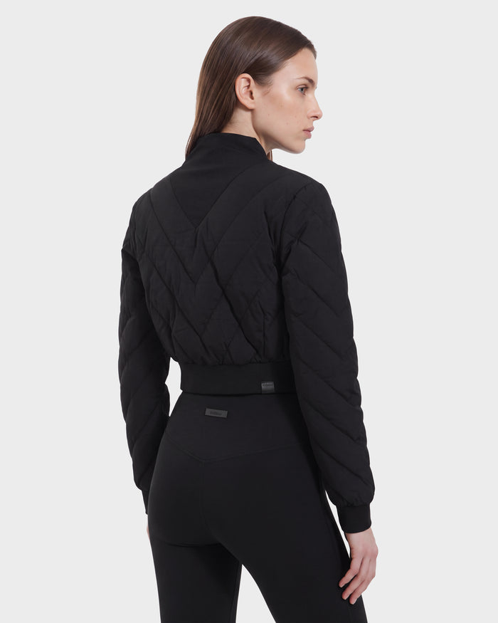 A woman with straight brown hair is seen from behind, looking to the right against a plain white background. She is wearing the Rudsak KATYA - 8526531 Black quilted jacket with down insulation, paired with black leggings.