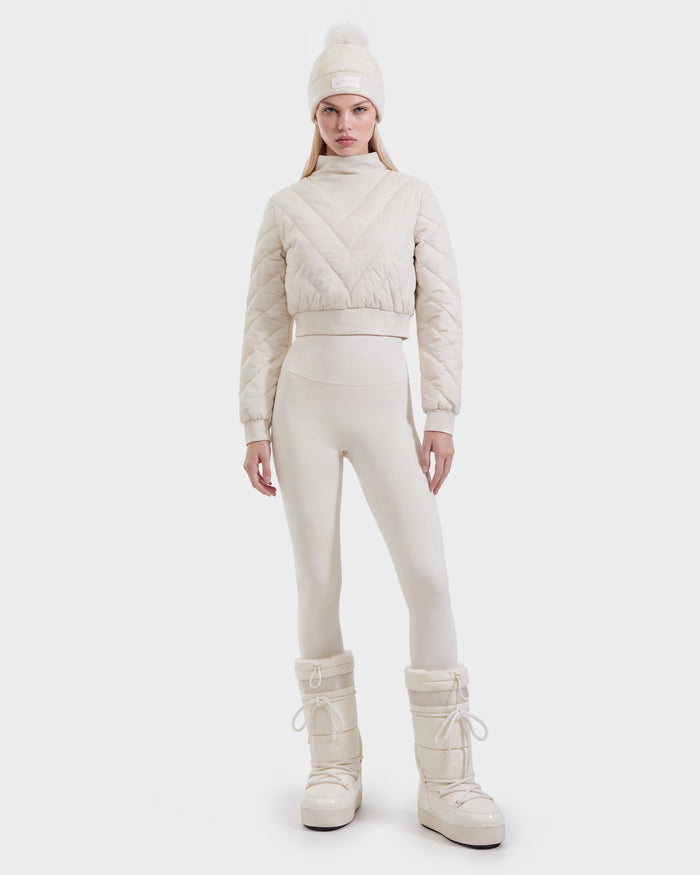A woman stands against a plain white background wearing the KATYA - 8526531 Vanilla quilted down jacket by Rudsak, paired with matching leggings, tall padded boots, and a beanie topped with a pom-pom.