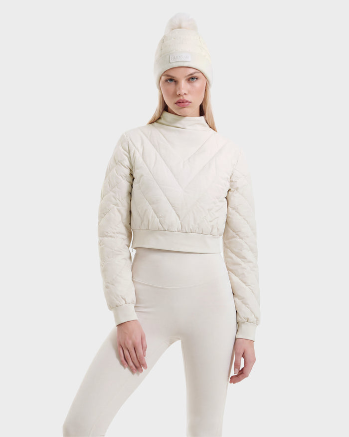 A woman wearing the Rudsak KATYA - 8526531 Vanilla cream quilted down jacket, paired with matching leggings and a knit beanie with a pom-pom, stands against a plain light background, looking at the camera.