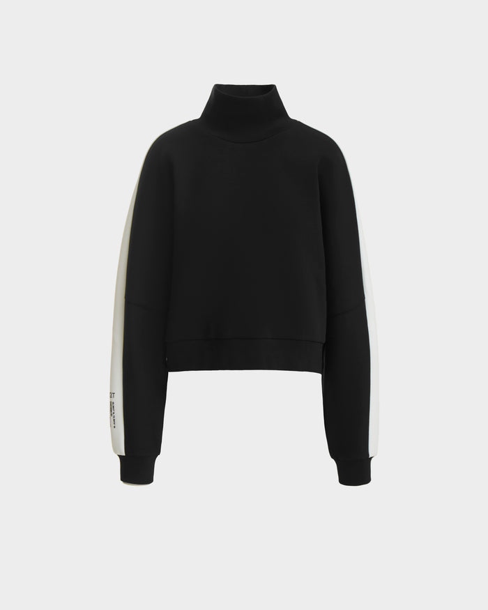 The KELLIE - 8524615 Black by Rudsak is a women’s long-sleeve mock neck top in black scuba fabric with white sleeve panels, offering a modern sporty style. Shown on a plain white background.