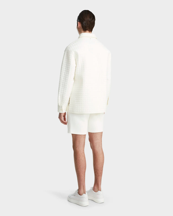 A person stands facing away, wearing the Rudsak KERRY - 9124003 Eggshell M shirt jacket with eco-friendly insulation, paired with white shorts and sneakers, set against a plain light background.