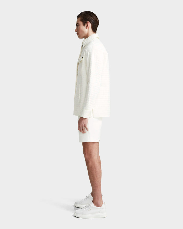 A person stands in profile against a plain light background, wearing the Rudsak KERRY - 9124003 Eggshell M unisex jacket with eco-friendly insulation, paired with white shorts and white platform sneakers.
