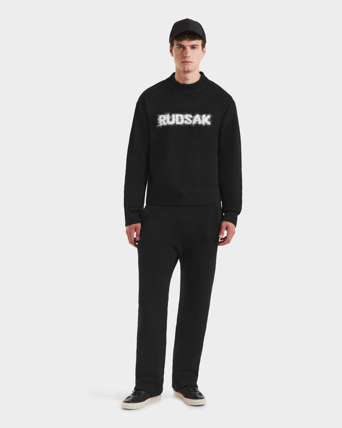 A man faces forward against a plain white background, wearing the Rudsak KINLEY - 9525555 Black M unisex crewneck sweater with "RUDSAK" in white, paired with black pants and shoes.
