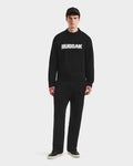 A man faces forward against a plain white background, wearing the Rudsak KINLEY - 9525555 Black M unisex crewneck sweater with "RUDSAK" in white, paired with black pants and shoes.