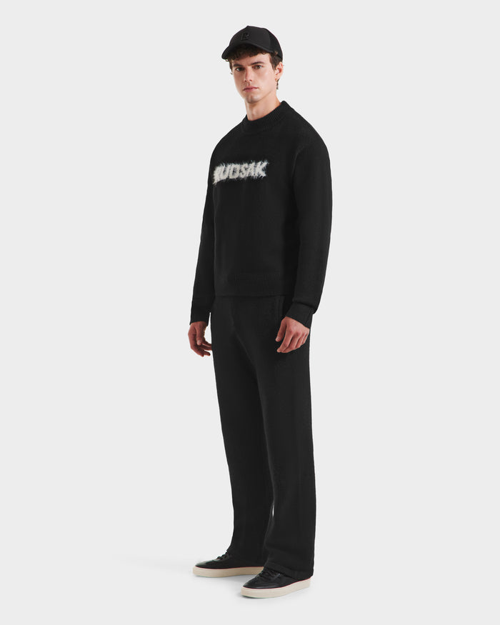 A man in the Rudsak KINLEY - 9525555 Black M textured knit sweater with a blurred white logo, wearing a black cap, black pants, and black sneakers stands against a plain white background.