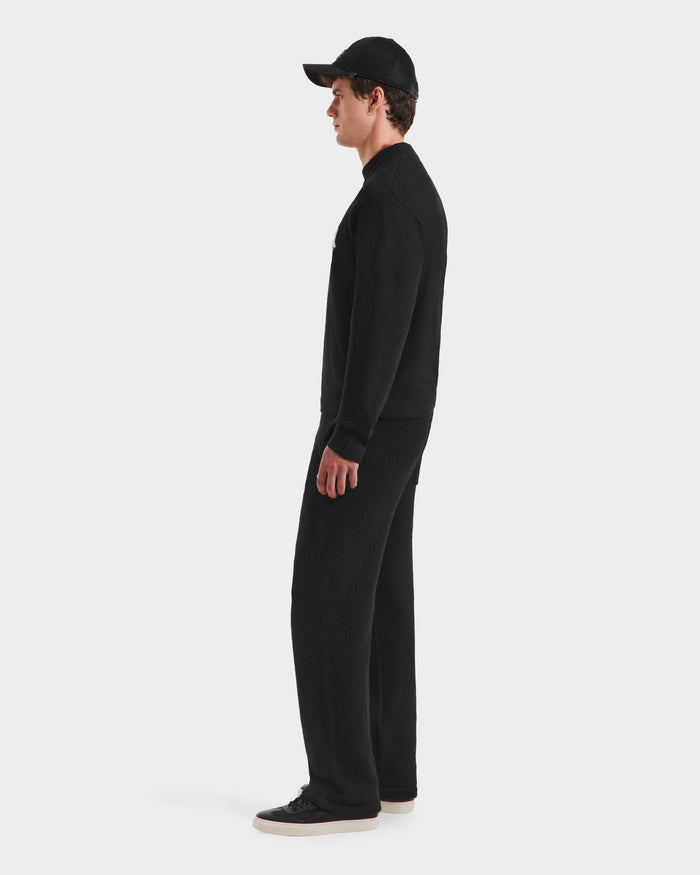 A person stands in profile against a white background, wearing the Rudsak KINLEY - 9525555 Black M sweater, along with a black cap, black pants, and black sneakers with white soles.