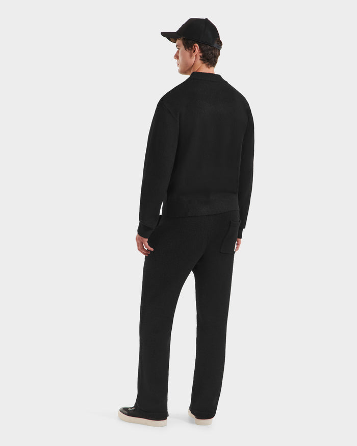 A man faces away wearing the Rudsak KINLEY - 9525555 Black M unisex crewneck sweater, black pants, beige-soled shoes, and a black cap, pictured against a plain white background.