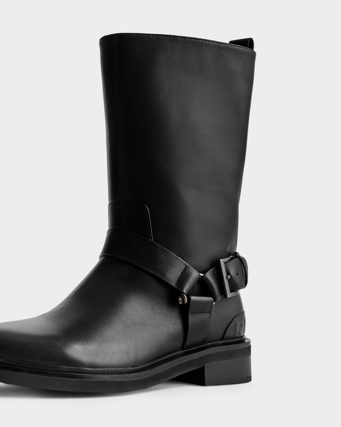 The KIRRA - 8225558 Black by Rudsak is a black leather mid-calf moto boot with a low heel, decorative ankle strap, and sleek side buckle detail, shown against a plain light background.