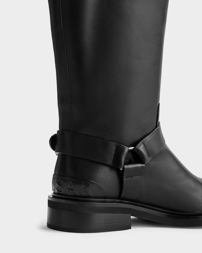 Close-up of the Rudsak KIRRA - 8225558 Black knee-high boot with black leather, low heel, ankle strap with metal ring, and embossed branding on the heel, set against a plain light background.