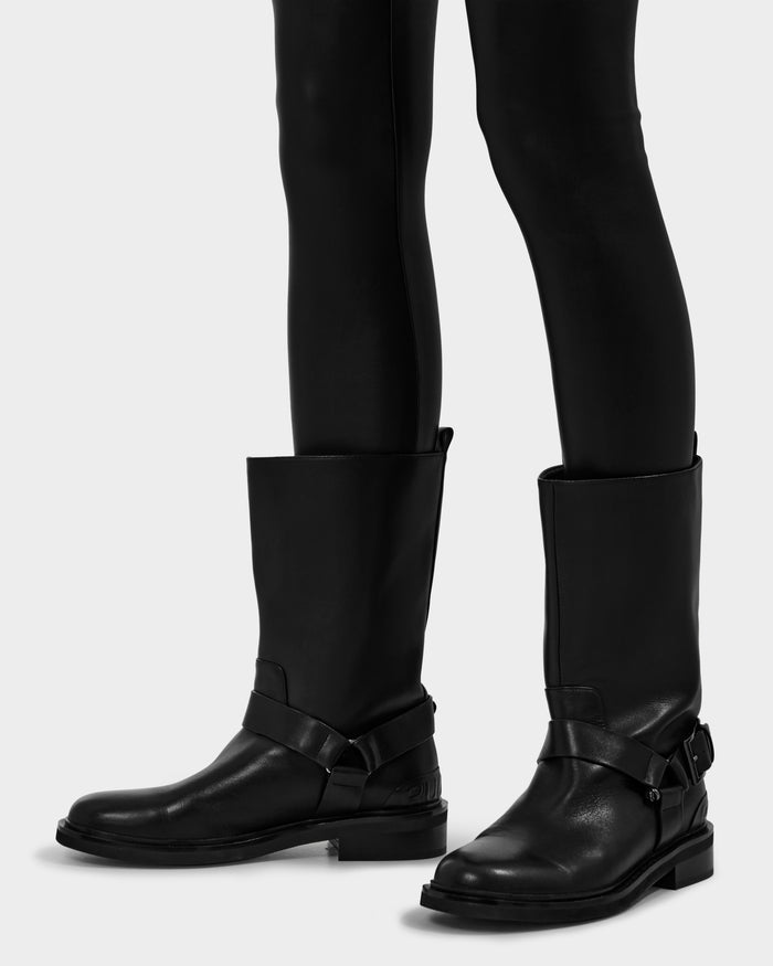 A person wears fitted black pants and KIRRA - 8225558 Black leather boots by Rudsak with harness details, standing on a light gray background.