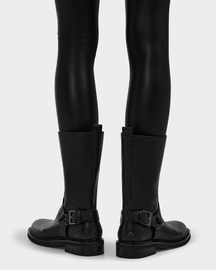 A person is shown from behind wearing Rudsak’s KIRRA - 8225558 Black leather moto boots with buckles, paired with shiny black leggings against a plain light background.