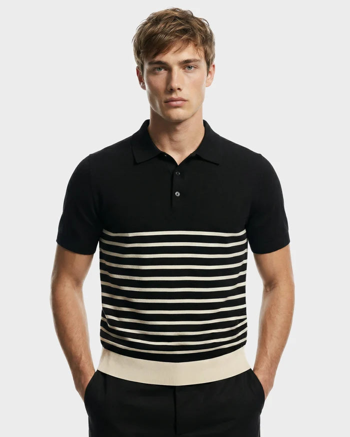 A young man with light brown hair models the KRIS - 6526005 Black men's designer polo by Rudsak, featuring black fabric with white horizontal stripes, standing hands-in-pockets against a plain light gray background.