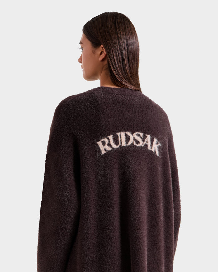 A woman with straight brown hair wears the Rudsak LACEY - 8525694 Espresso dark fuzzy coat, perfect for cozy layering, featuring "RUDSAK" in white on the back, as she stands facing left against a plain light background.