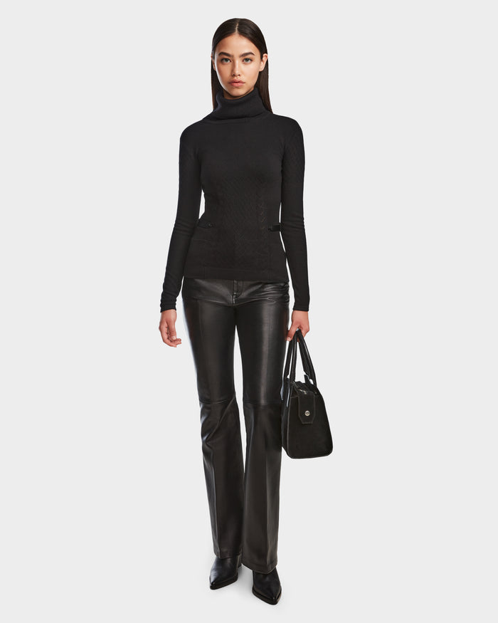 A woman faces forward against a plain white background, wearing the Rudsak LAURA - 8524596 Black womens knit turtleneck sweater, black leather pants with vegan leather trim, pointed-toe shoes, and holding a black handbag in her left hand.