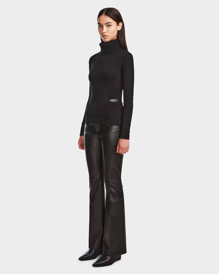 A woman with straight dark hair and a neutral expression stands against a plain background wearing the Rudsak LAURA - 8524596 Black leather flared pants with vegan leather trim, paired with a knit turtleneck sweater and black heeled boots.