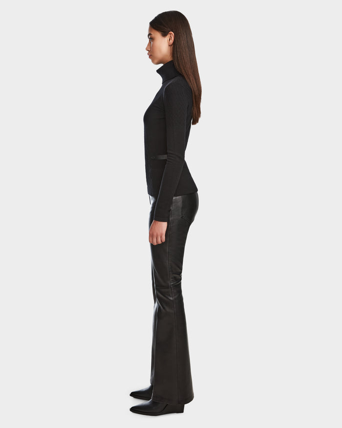 A woman in profile wears the LAURA - 8524596 Black knit turtleneck sweater by Rudsak with black flared leather pants and heeled boots, standing against a plain white background.