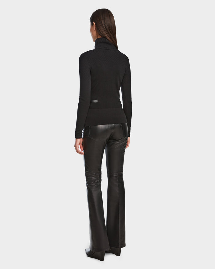 A woman with straight brown hair is standing and facing away, wearing the Rudsak LAURA - 8524596 Black womens knit turtleneck sweater with black flared leather pants and pointed-toe shoes against a plain white background.