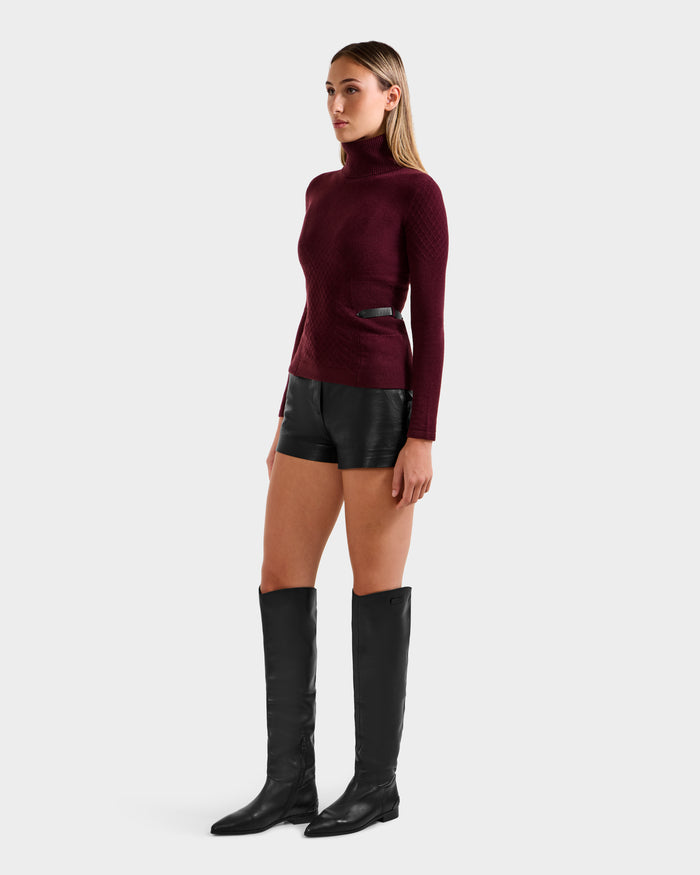 A woman faces left in a Rudsak LAURA - 8524596 Bordeaux women's knit turtleneck sweater, black leather shorts with vegan trim, and tall black boots against a plain light background.