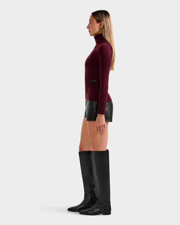 A woman stands in profile wearing the Rudsak LAURA - 8524596 Bordeaux women’s knit turtleneck sweater, paired with black leather shorts and tall black boots against a plain light background.