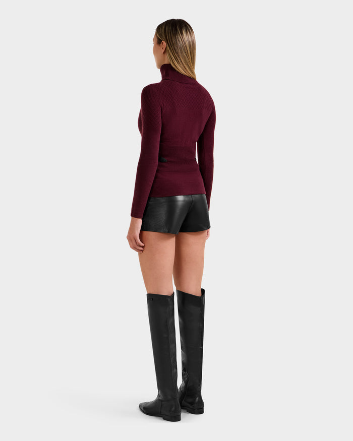 A woman stands facing away, wearing the Rudsak LAURA - 8524596 Bordeaux women's knit turtleneck sweater, black shorts with vegan leather trim, and tall black boots against a plain light background.