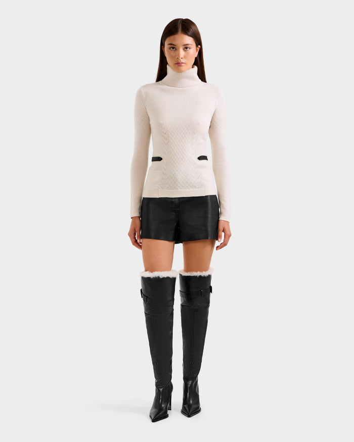 A woman faces forward wearing the Rudsak LAURA - 8524596 Vanilla knit turtleneck sweater, black high-waisted shorts, and black pointed-toe over-the-knee boots against a plain light gray background.