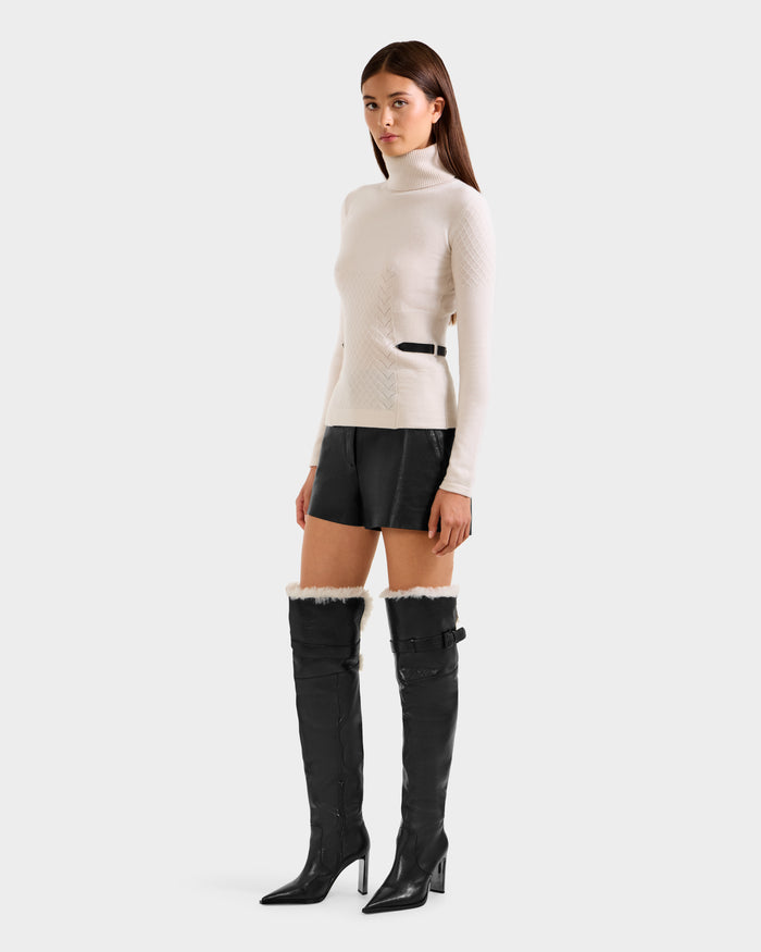 A woman stands against a plain background wearing the Rudsak LAURA - 8524596 Vanilla women's knit turtleneck sweater, paired with black leather shorts and tall black heeled boots featuring fur trim and buckle details.