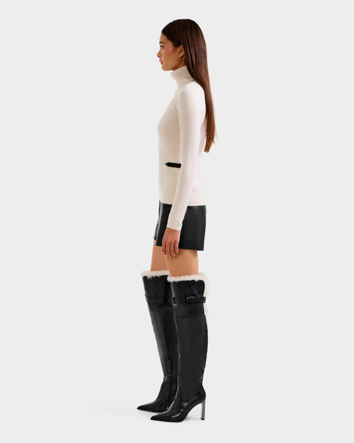 A woman in profile models the Rudsak LAURA - 8524596 Vanilla women’s knit turtleneck sweater, styled with black leather shorts and high-heeled knee-high boots featuring vegan leather trim, against a white background.
