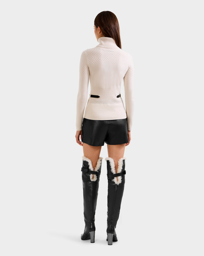 A woman faces away, wearing the Rudsak LAURA - 8524596 Vanilla women's knit turtleneck sweater with black shorts and black knee-high heeled boots with white fur lining, set against a plain light background.
