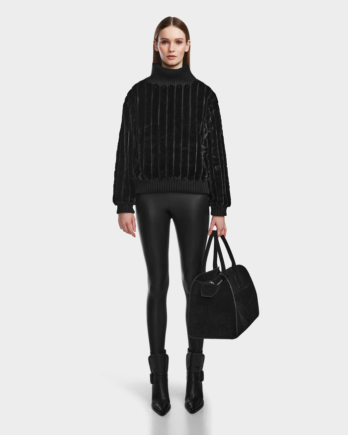 A woman stands facing forward in the LAYLA - 8524610 Black turtleneck by Rudsak, paired with shiny black leggings, black heeled boots, and a large black handbag against a white background.