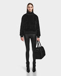 A woman stands facing forward in the LAYLA - 8524610 Black turtleneck by Rudsak, paired with shiny black leggings, black heeled boots, and a large black handbag against a white background.