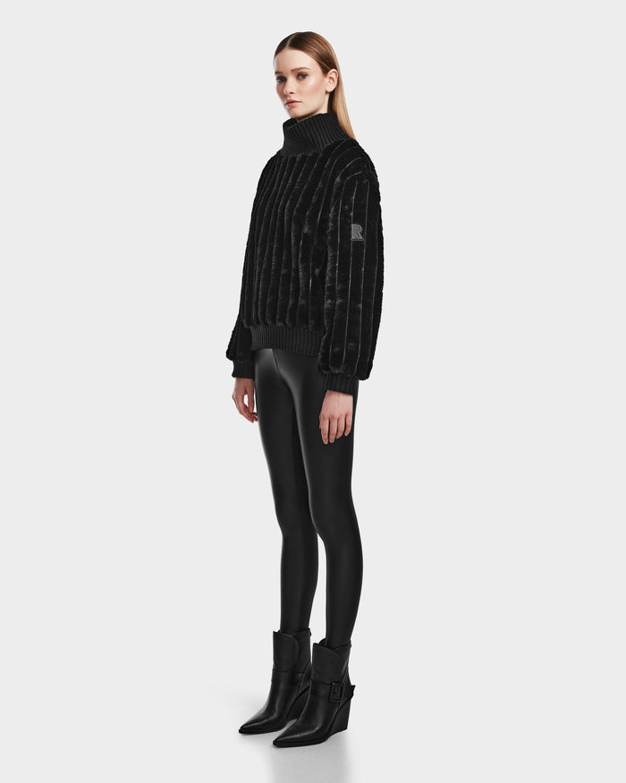 A woman stands against a plain background wearing the LAYLA - 8524610 Black turtleneck sweater by Rudsak, shiny black leggings, and black ankle boots. She faces slightly to the side with her arms relaxed at her sides.