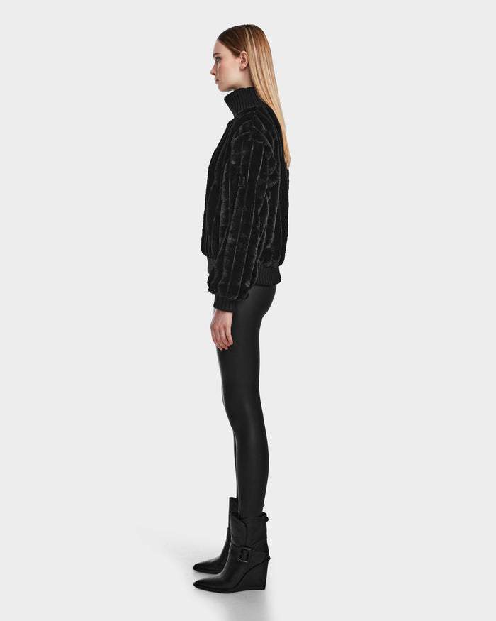 A woman with straight blonde hair stands in profile, wearing the LAYLA - 8524610 Black faux fur turtleneck by Rudsak, paired with black fitted pants and buckled ankle boots, against a plain white background.
