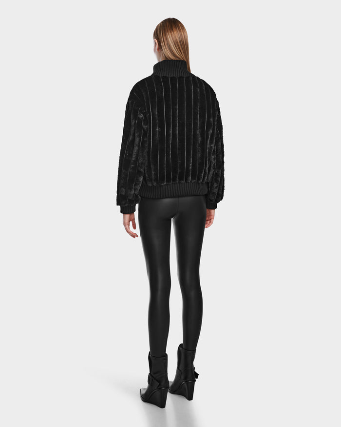 A person with straight, light brown hair faces away, wearing the Rudsak LAYLA - 8524610 Black turtleneck, shiny black leggings, and black ankle boots against a plain light background.