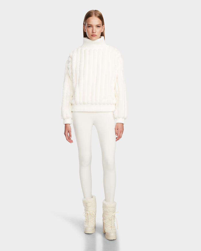 A woman with straight, light brown hair and a neutral expression models the LAYLA - 8524610 Iceland turtleneck sweater by Rudsak, paired with white fitted pants and winter boots against a plain background.