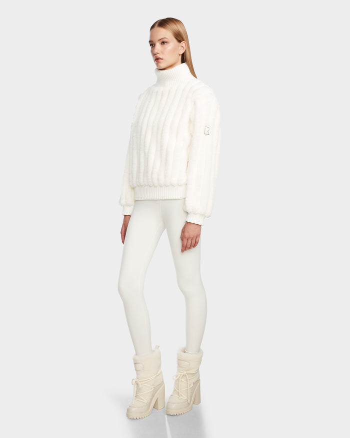A woman poses against a light gray background in the LAYLA - 8524610 Iceland turtleneck by Rudsak, styled with white leggings and cream winter boots. Her straight blonde hair and neutral expression complete the chic look.
