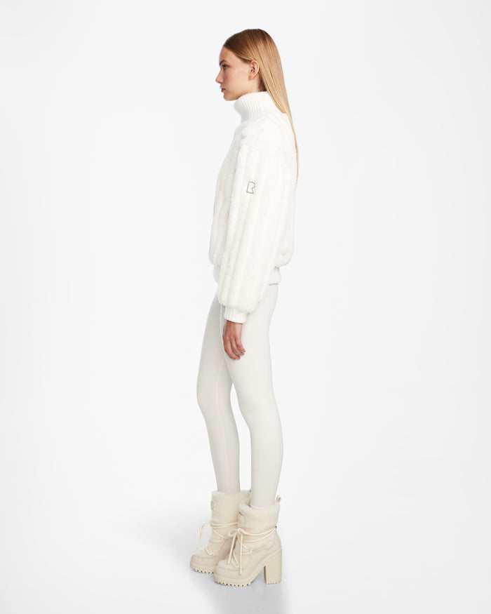 A woman with straight blonde hair stands in profile against a white background, wearing the LAYLA - 8524610 Iceland faux fur turtleneck by Rudsak, fitted white pants, and white platform boots with fur trim.
